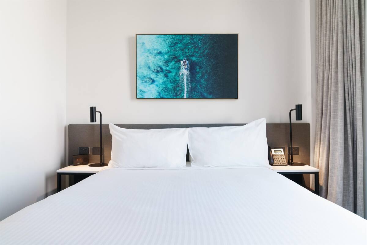 A hotel room with a large bed, two bedside tables, a nightstand, a telephone, and a painting of a ship on a blue background.