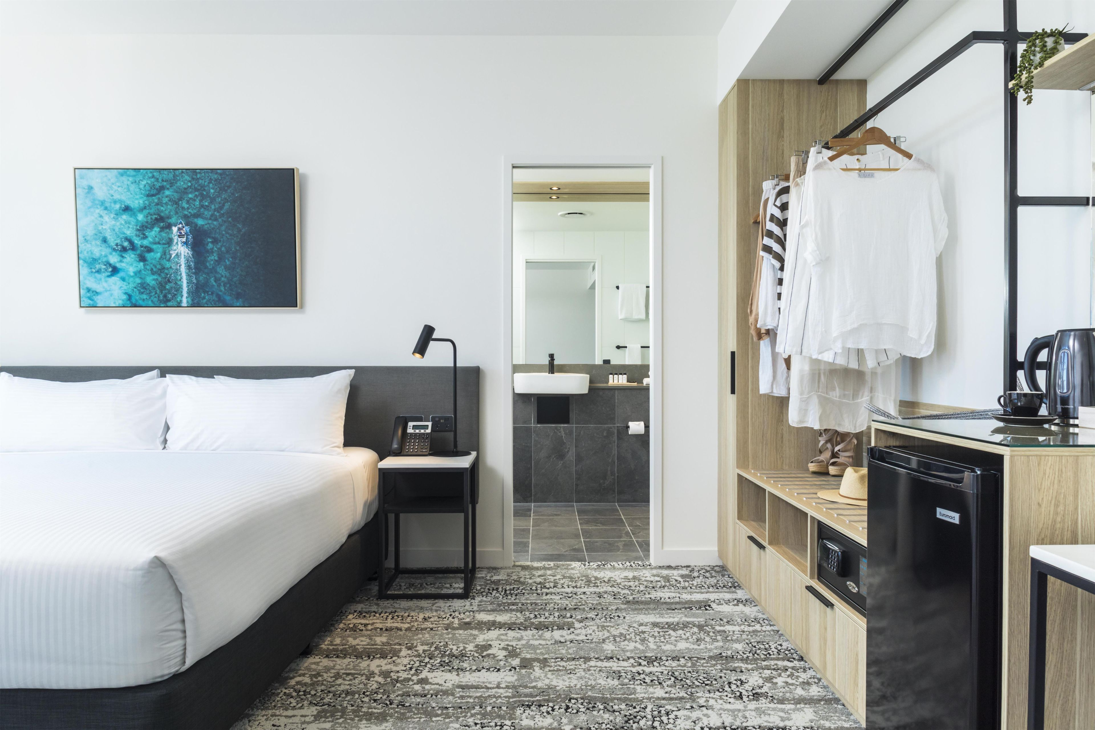 A hotel room with a bed, a desk with a telephone, a mirror, a bathroom visible through an open door, and a kitchenette area.