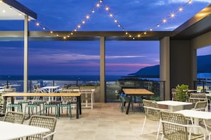 Un patio al aire libre con mesas y sillas, luces colgantes y vista al océano.