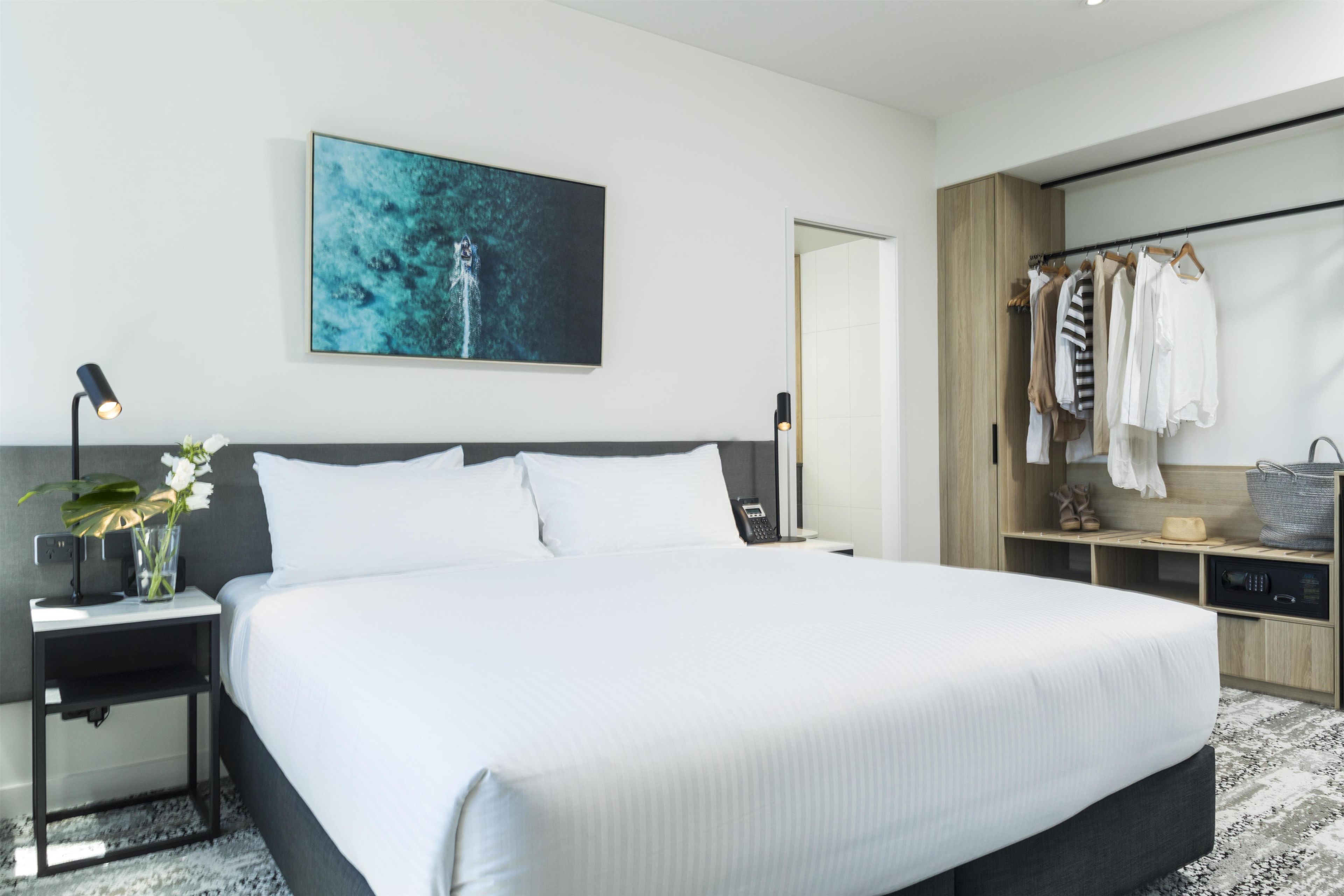 A modern hotel room with a large bed, a bedside table with a lamp, a painting on the wall, and a wardrobe with hanging clothes.
