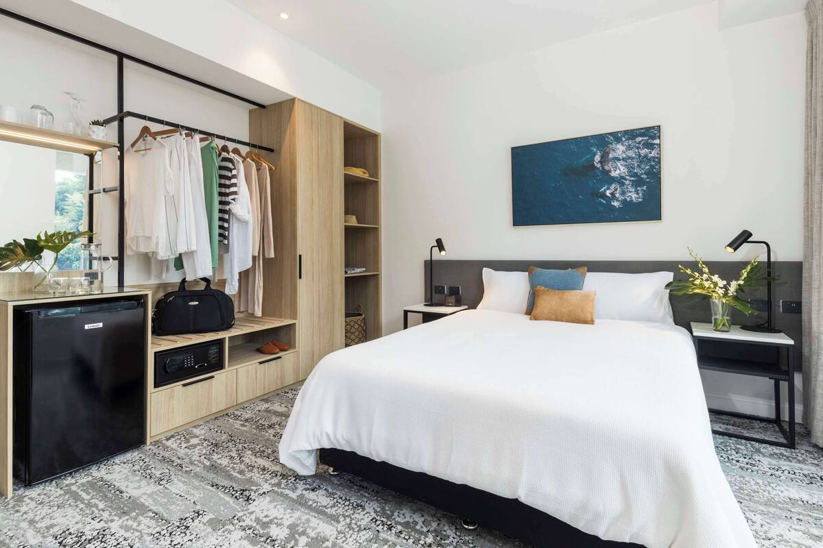 A modern hotel room with a large bed, a wardrobe, a desk with a computer, and a small refrigerator.