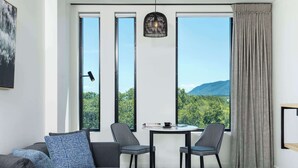 Una habitación moderna con un sofá gris, dos sillas, una mesita y vistas a zonas verdes y montañas a través de grandes ventanales.