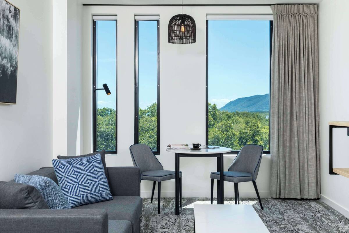 A modern room with a gray sofa, two chairs, a small table, and a view of greenery and mountains through large windows.