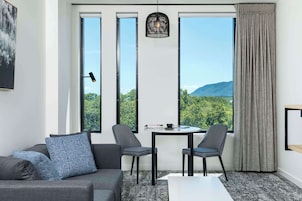Une pièce moderne avec un canapé gris, deux fauteuils, une petite table et une vue sur la verdure et les montagnes, grâce à de grandes fenêtres.