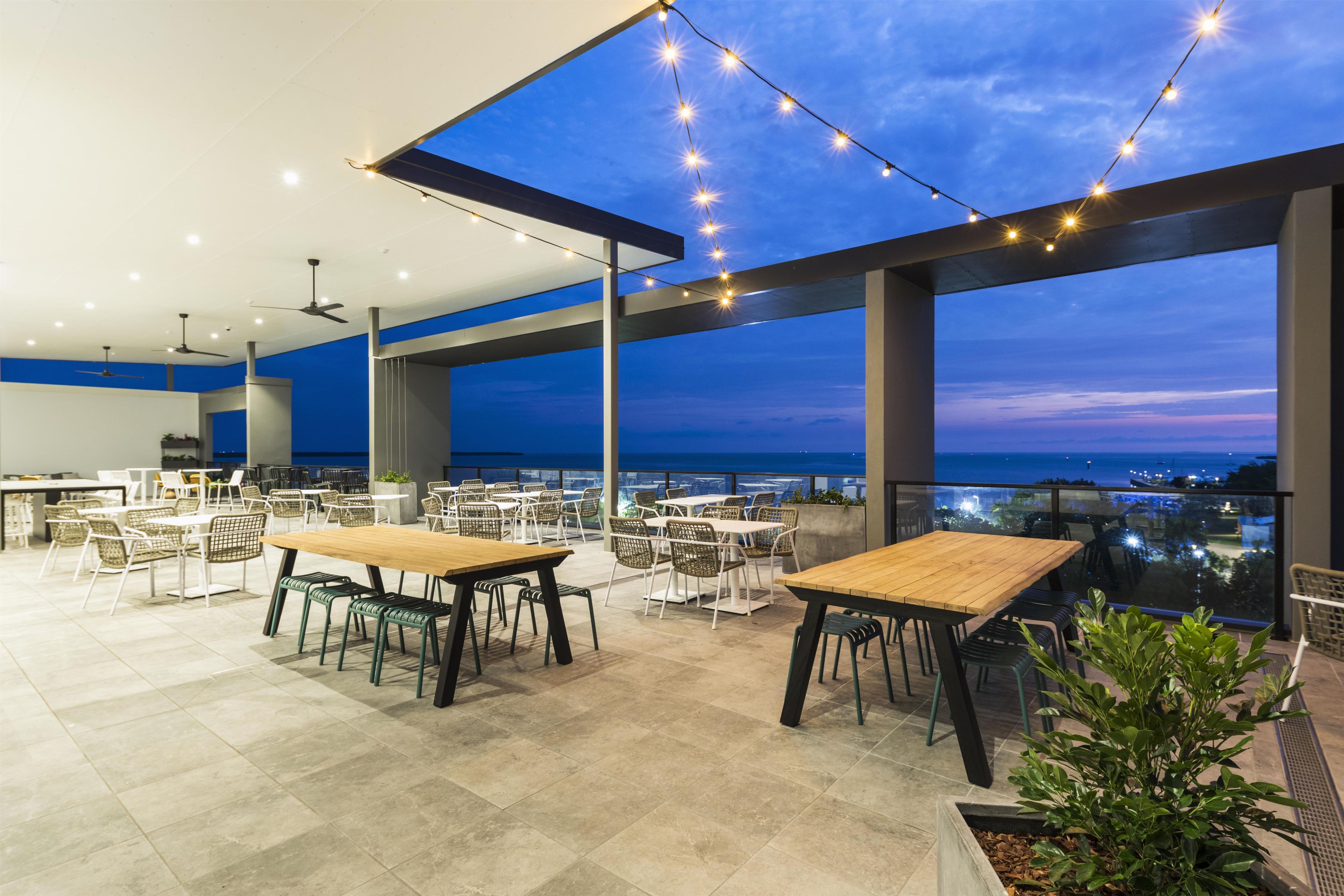 Un espacio exterior para comer con mesas y sillas, luces colgantes y vistas del paisaje urbano al anochecer.
