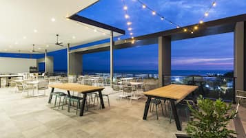 An outdoor dining area with tables and chairs, string lights, and a view of the cityscape at dusk.