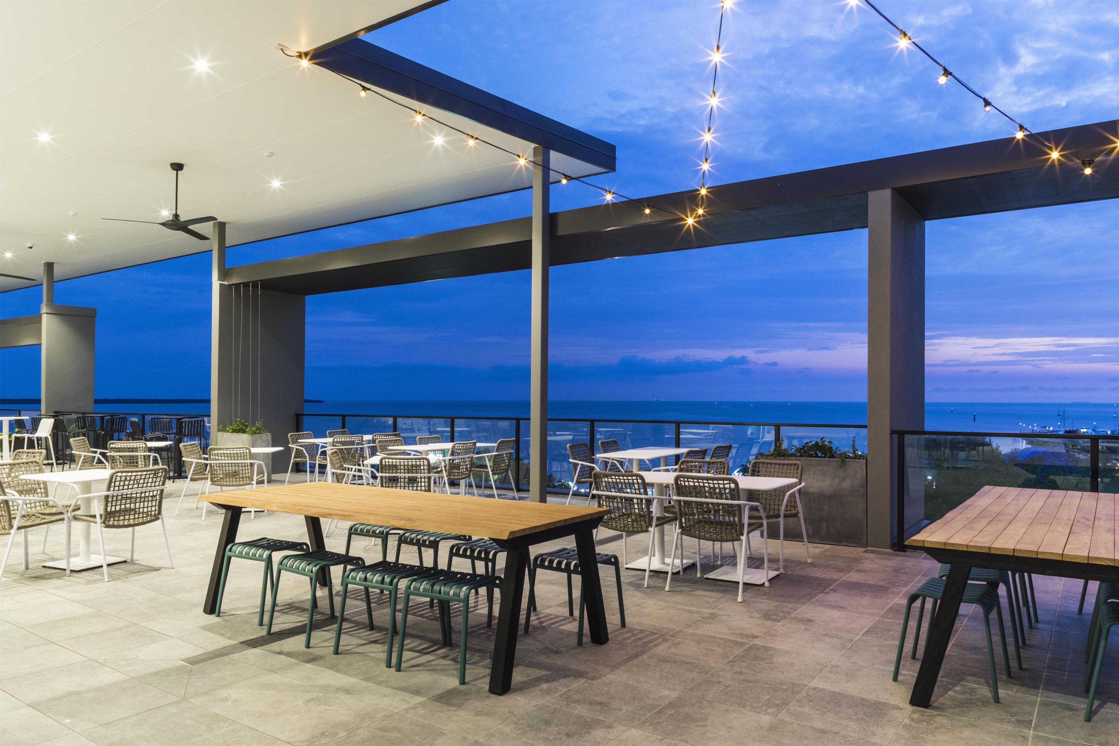 An outdoor dining area with tables and chairs, string lights, and a view of the sea.