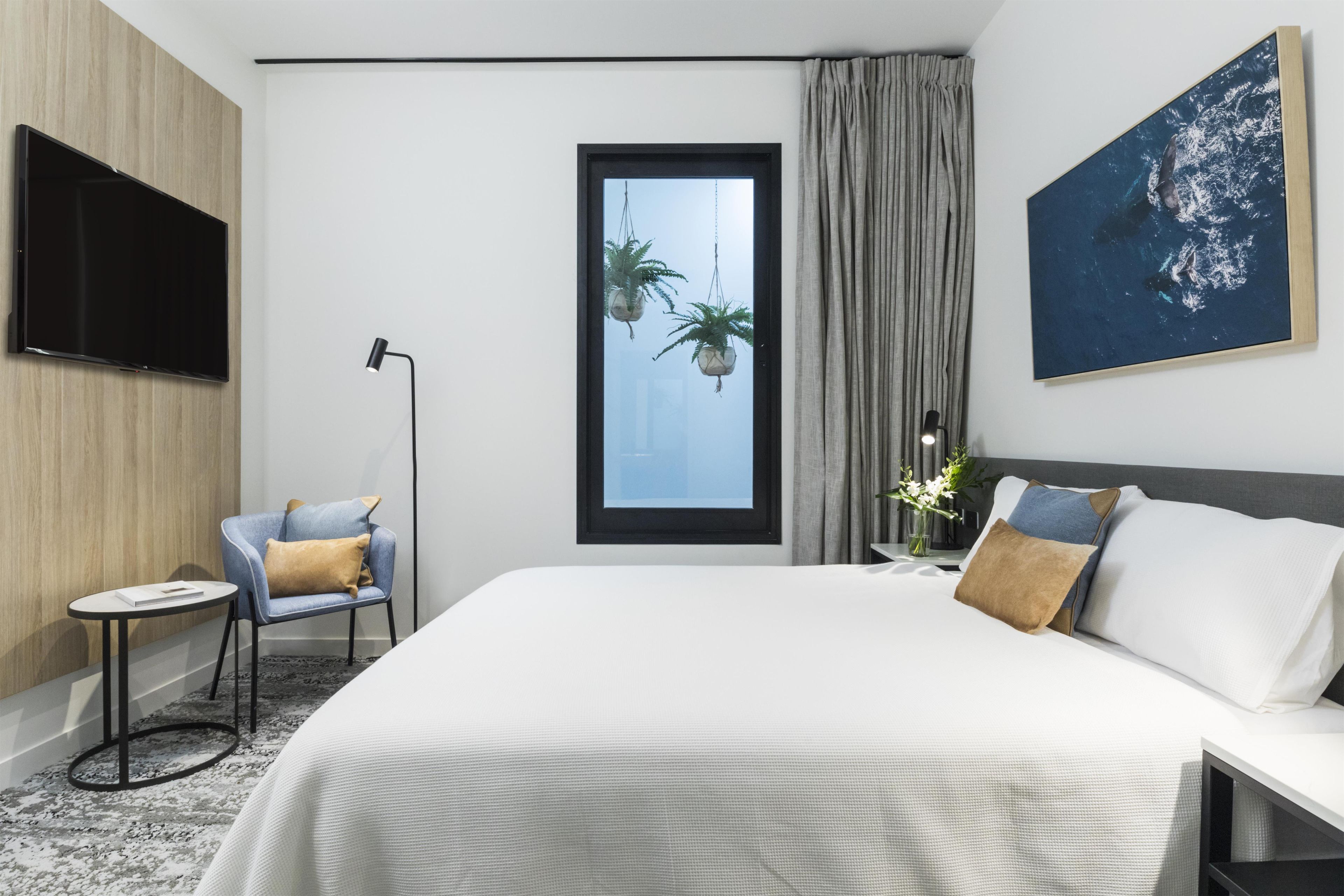 A hotel room with a bed, a chair, a TV, a window with curtains, and a painting.