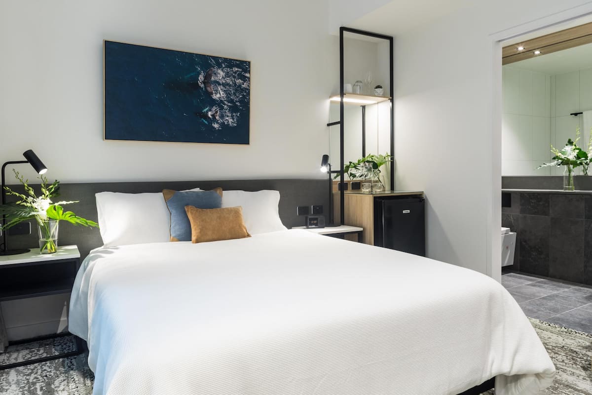 A modern hotel room with a large bed, a bedside table with a lamp, a small cabinet, and a large mirror.