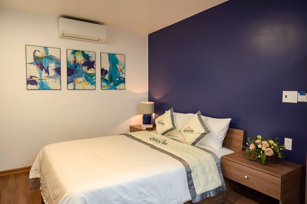 A neatly made bed with a white bedspread and pillows, a bedside table with a lamp, and a wall with abstract art.