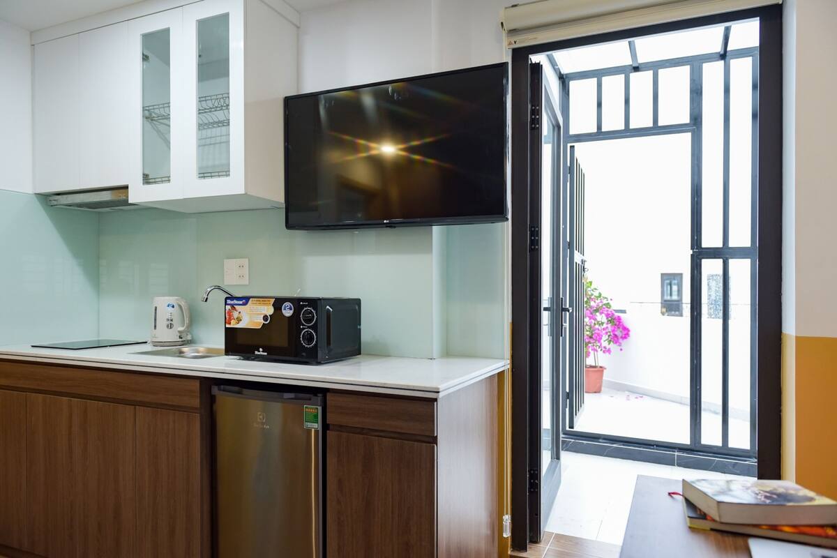 A modern kitchen with a built-in microwave, a kettle, and a dishwasher. A flat-screen TV is mounted above the counter, and a sliding glass door leads to an outdoor area with a potted plant.