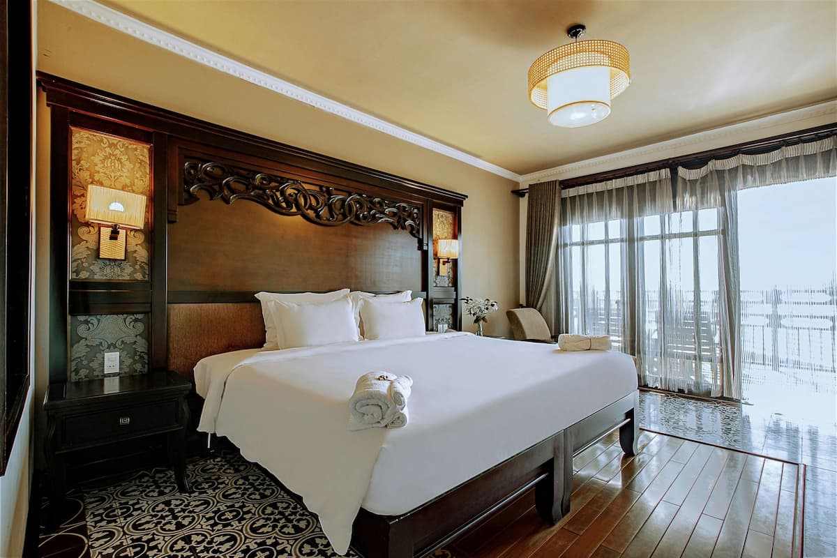 A large bed with a wooden headboard, two bedside lamps, and a rolled towel on the bed.