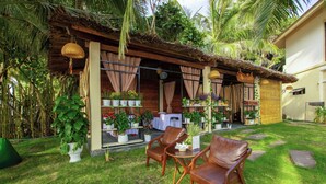 A tropical outdoor seating area with wooden chairs and a table, surrounded by potted plants and a thatched roof structure. - Rainbow Apartment Hai Phong (Hai Phong)
