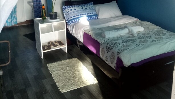 A bedroom with a bed, a nightstand, a small shelf, a potted plant, and a hanging light fixture. - Firebird Inn (Naivasha)