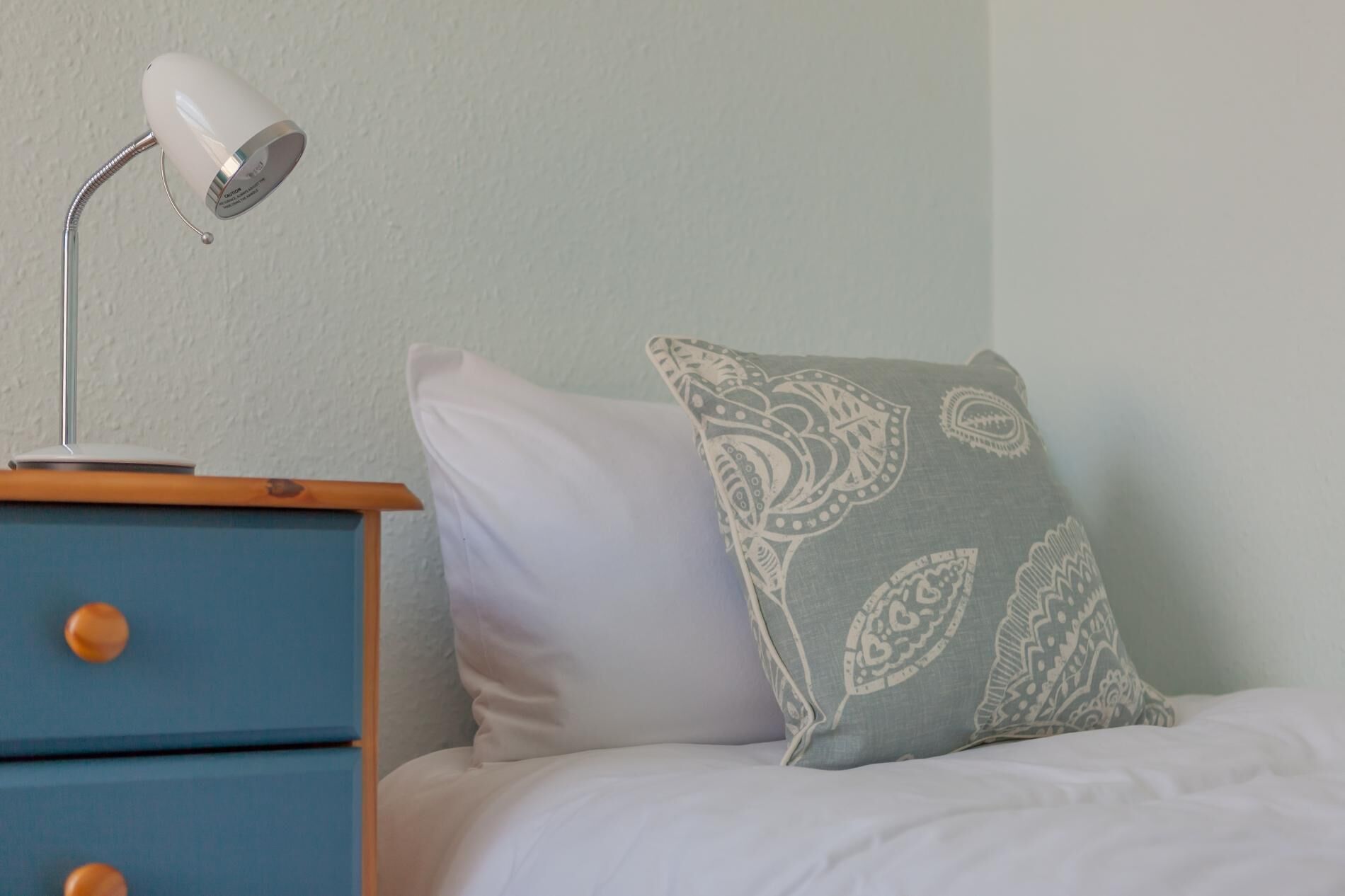 A bed with a white pillow and a patterned pillow, a blue nightstand with a white lamp, and a plain wall.