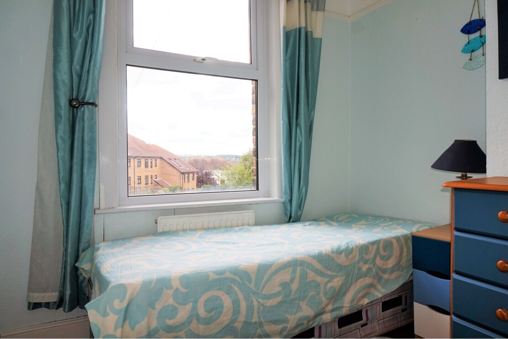 A bedroom with a single bed, a dresser, a window with curtains, and a view of buildings outside.