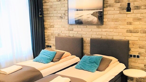 A modern hotel room with two beds, a brick wall, a large window with curtains, a small round table, and a picture on the wall.