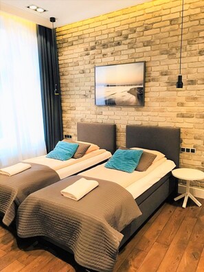 A modern hotel room with two beds, a brick wall, a large window with curtains, a small round table, and a picture on the wall. - Victus Apartamenty - Omega (Sopot)