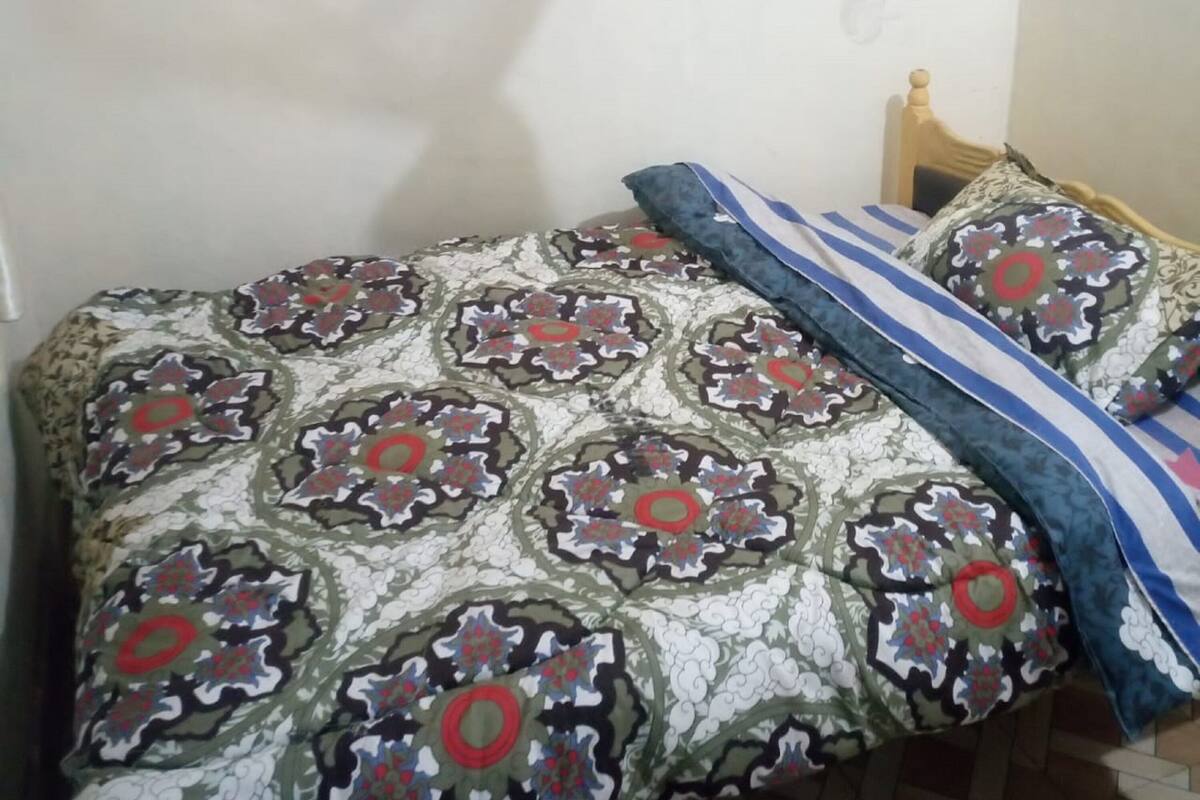 Uma cama com colcha estampada e almofadas, cabeceira de madeira e luminária fixada na parede.