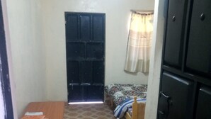 A small room with a tiled floor, a bed with a patterned bedspread, a wooden table with folded items, and a black wardrobe.