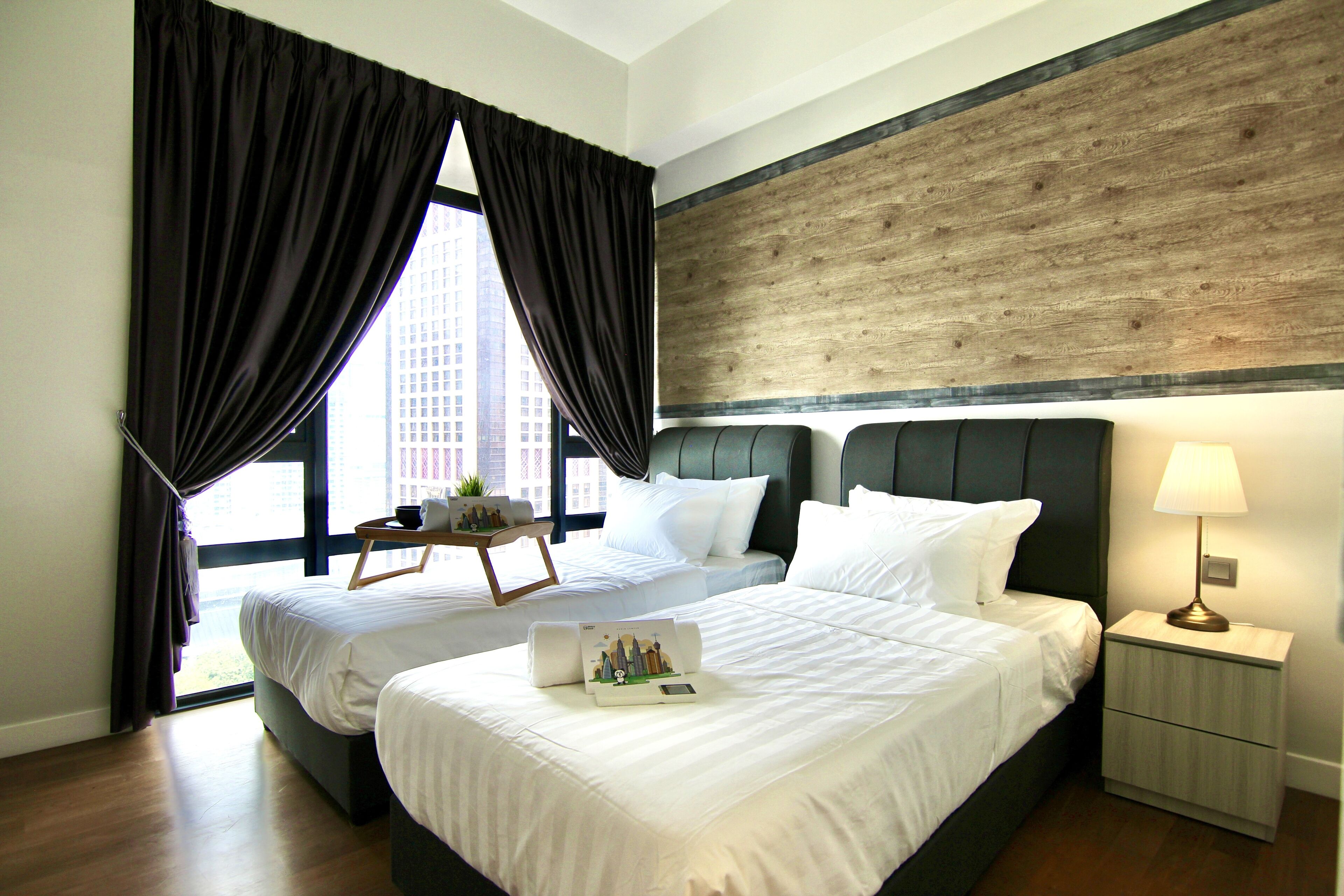 Habitación de hotel con dos camas, cabecero de madera, mesita de noche con lámpara y vistas a la ciudad a través de amplias ventanas.