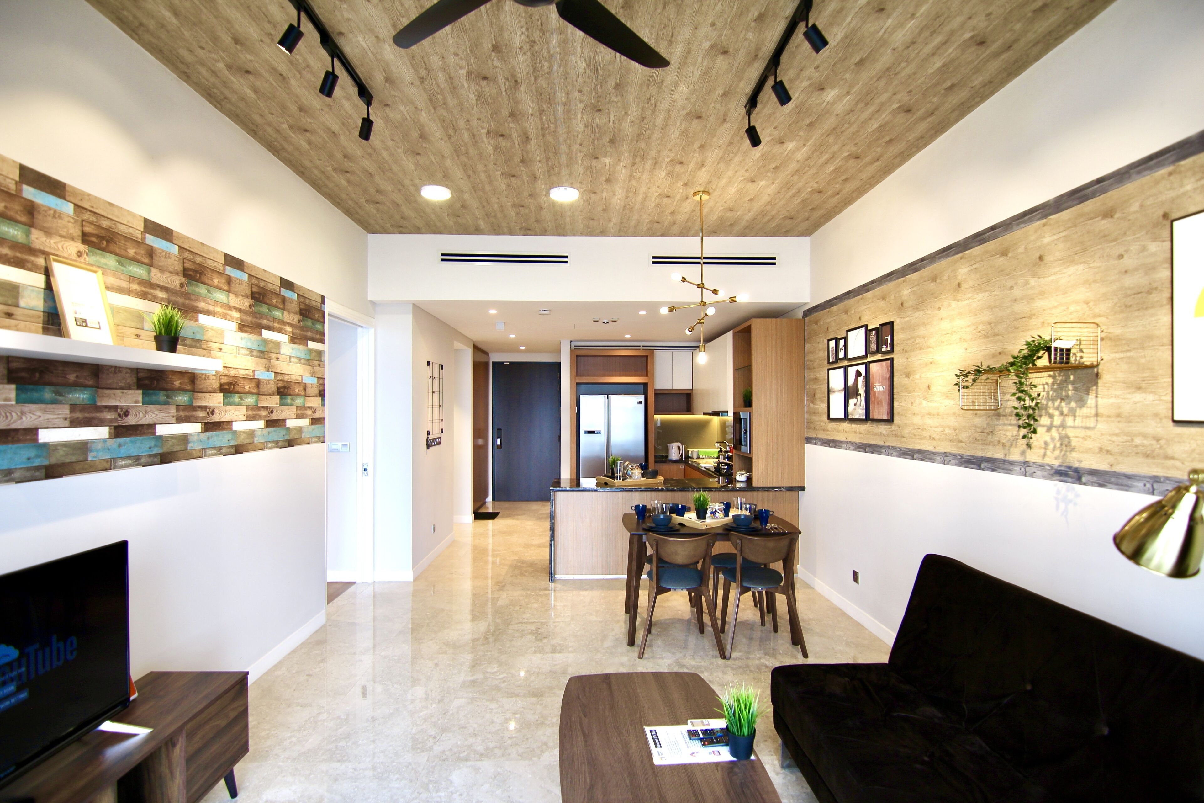 A modern living room with a wooden ceiling, a flat-screen TV, a dining area with a table and chairs, and a kitchen in the background.