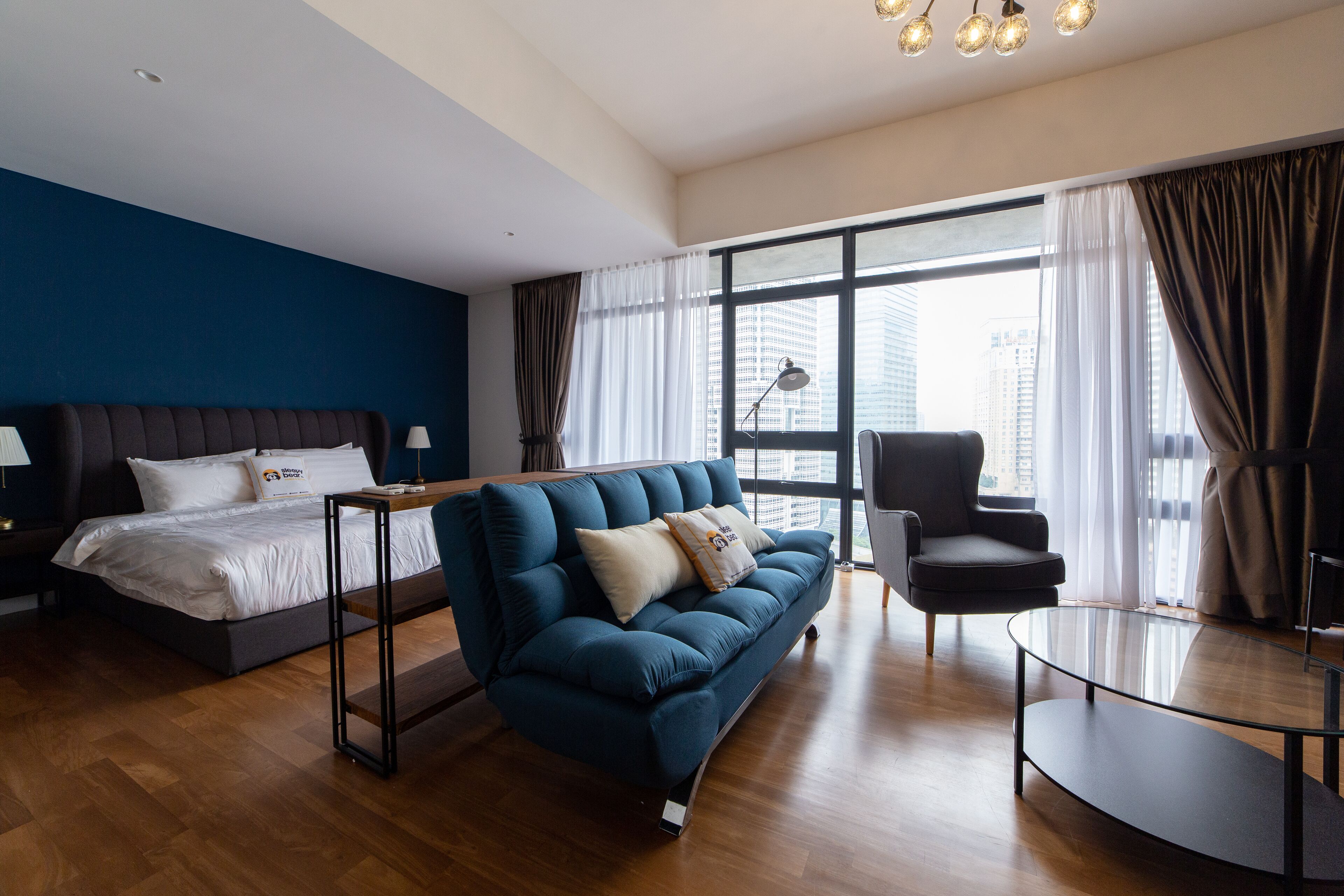 Habitación de hotel moderna con una cama grande, un sofá azul, una mesa de centro de vidrio y vistas a la ciudad a través de amplias ventanas.