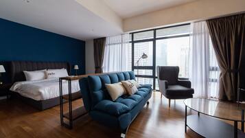Habitación de hotel moderna con una cama grande, un sofá azul, una mesa de centro de vidrio y vistas a la ciudad a través de amplias ventanas.