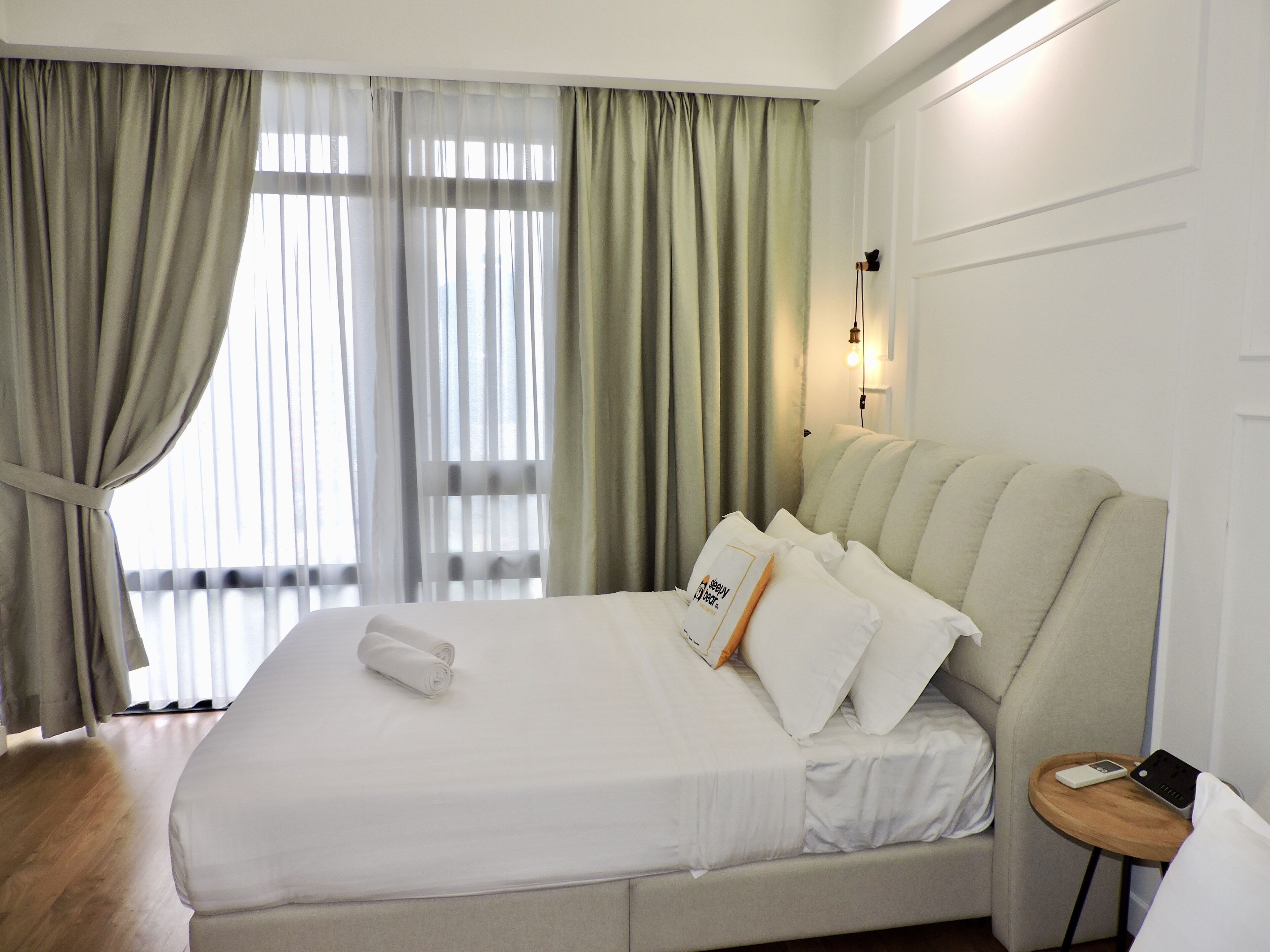 A neatly made bed with pillows and a rolled towel, a bedside table with a phone and a small device, and a window with sheer curtains.