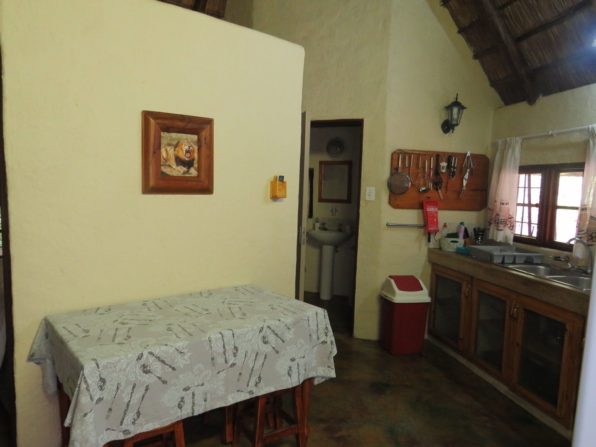 A small kitchen with a sink, a table with a tablecloth, a framed picture on the wall, and a window with curtains.