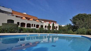 A swimming pool with a clear blue water surface, surrounded by a well-maintained hedge and a building with a tiled roof and arched windows. - 3-room apartment with large private garden, air conditioning and parking (Cavalaire-sur-Mer)