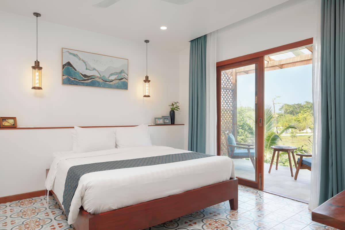 A neatly made bed with a patterned bedspread, a painting on the wall, and a view of the outdoors through a sliding glass door.