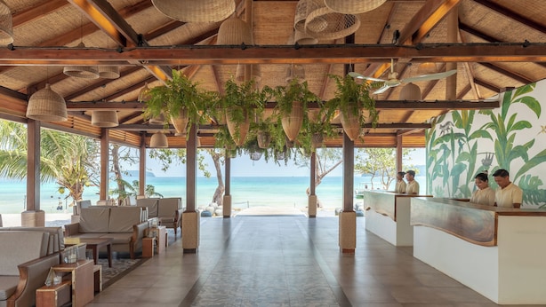 A beachside reception area with a thatched roof, hanging planters, and a view of the sea.