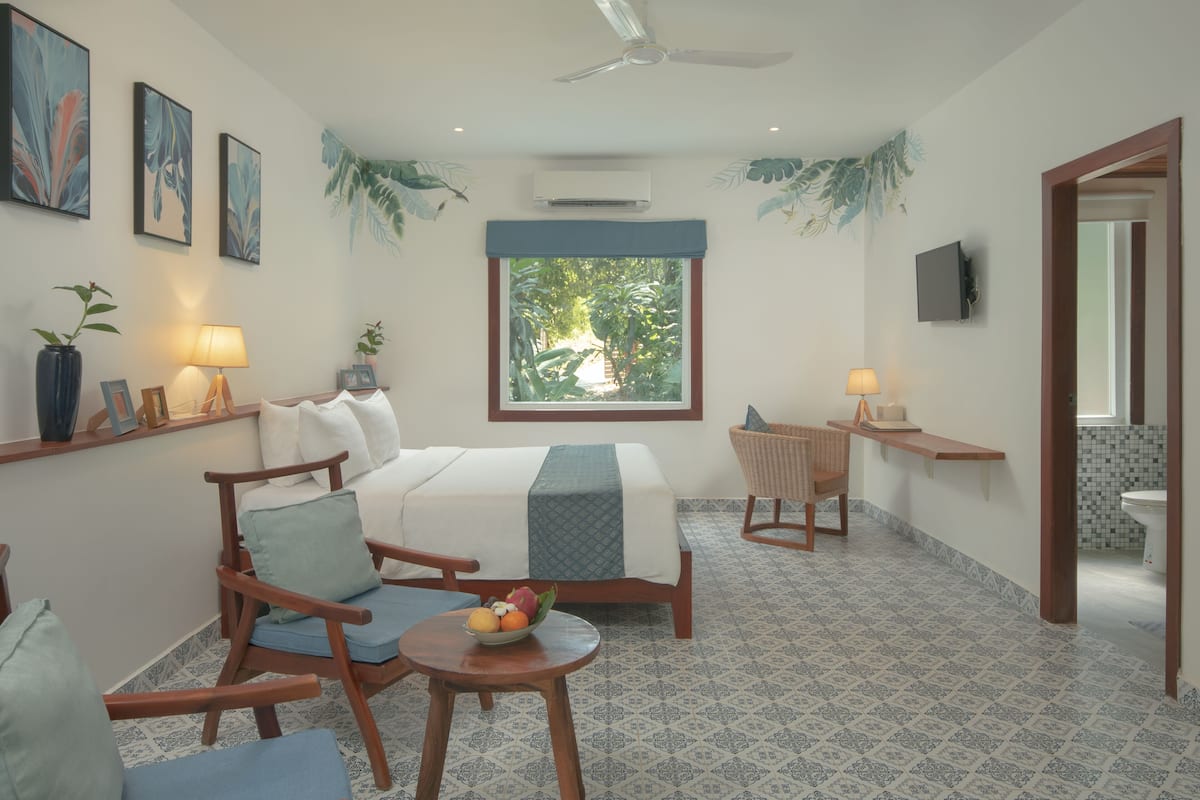 A bedroom with a bed, two chairs, a small table with fruit, a window with a view of greenery, and a TV on the wall.
