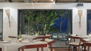 A restaurant with wooden tables and chairs, white tablecloths, and a view of the beach and trees through large windows.