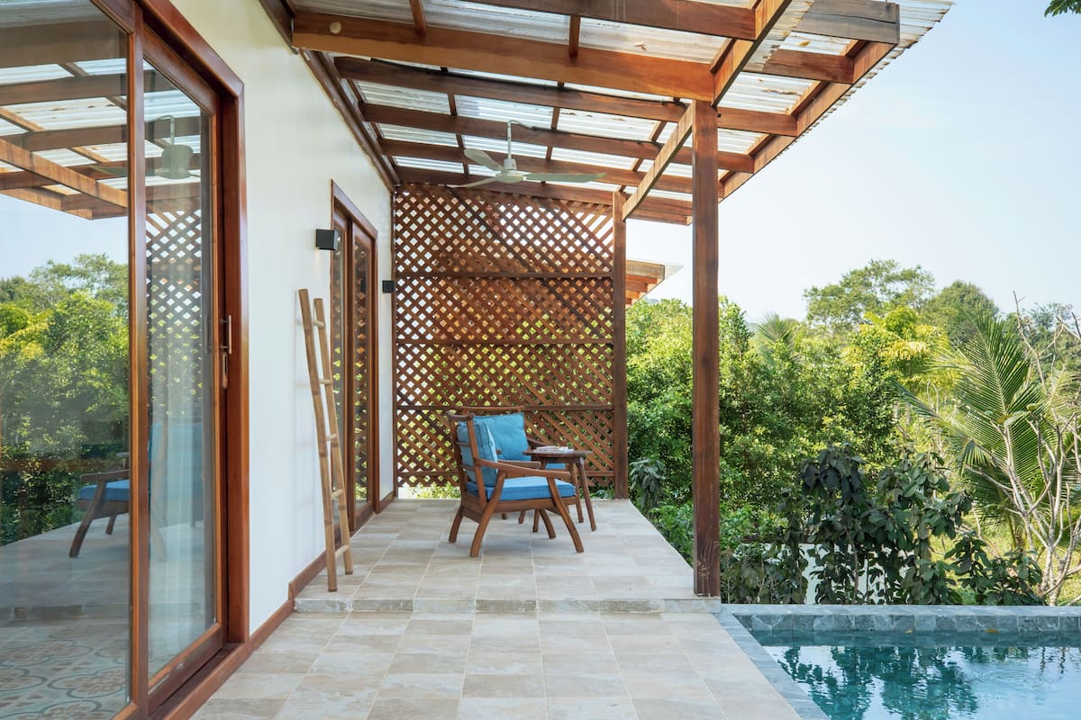 A covered outdoor seating area with a pool, wooden furniture, and a view of lush greenery.