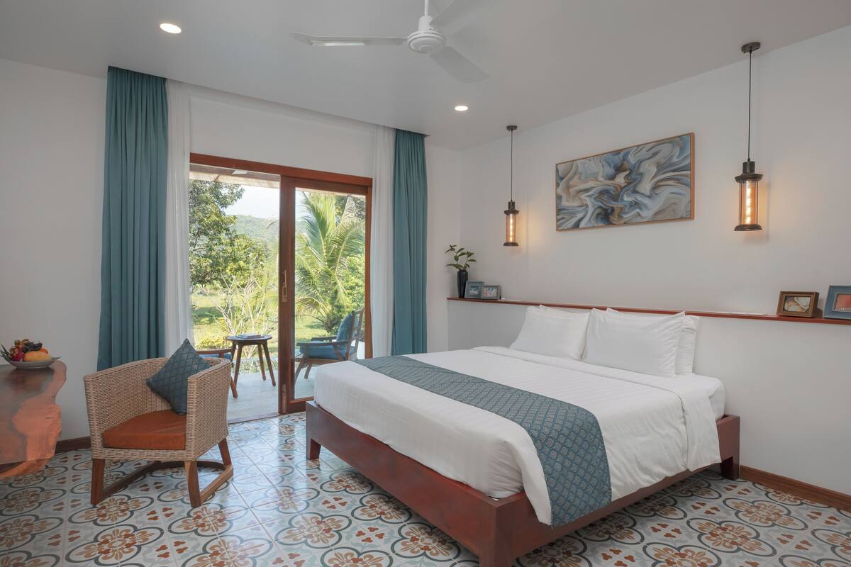 A bedroom with a bed, a chair, a ceiling fan, a window with curtains, and a wall-mounted light fixture.