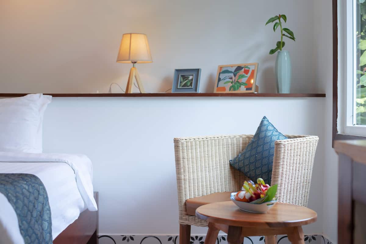A hotel room with a bed, a wicker chair with a bowl of fruit, a lamp, and framed pictures on the wall.