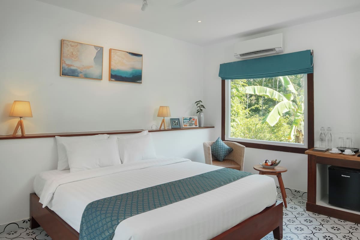 A neatly made bed with a patterned bedspread, a bedside table with a lamp, a small seating area with a cushion, and a window with a view of greenery.