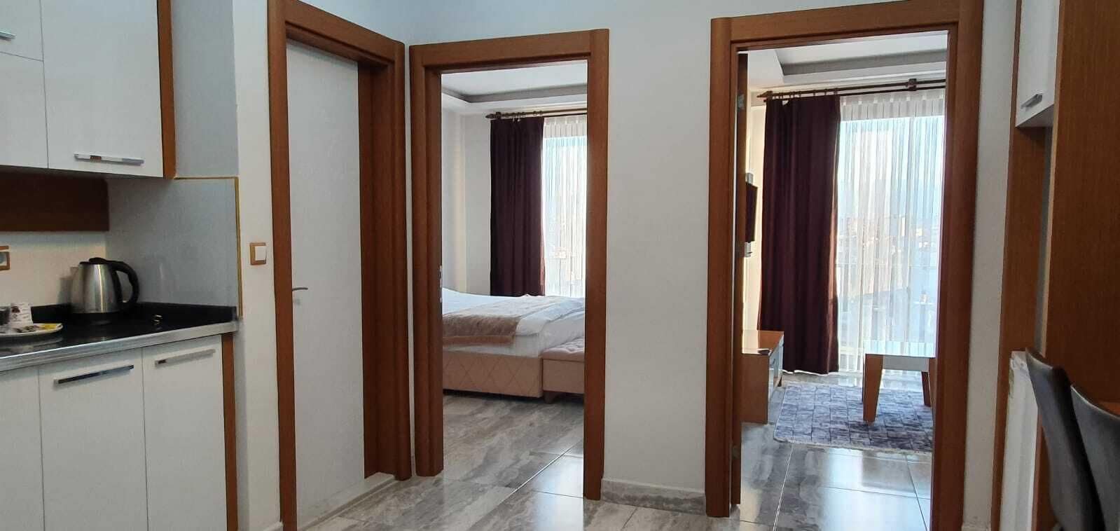 Un pasillo estrecho que lleva a un dormitorio con cama y ventana con cortinas. A la izquierda, hay una zona de kitchenette con armarios y hervidor.