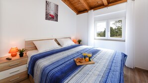 Een slaapkamer met een houten plafond, een bed met een blauw-witte strepen deken, een nachtkastje met een lamp en een raam met uitzicht op groen.
