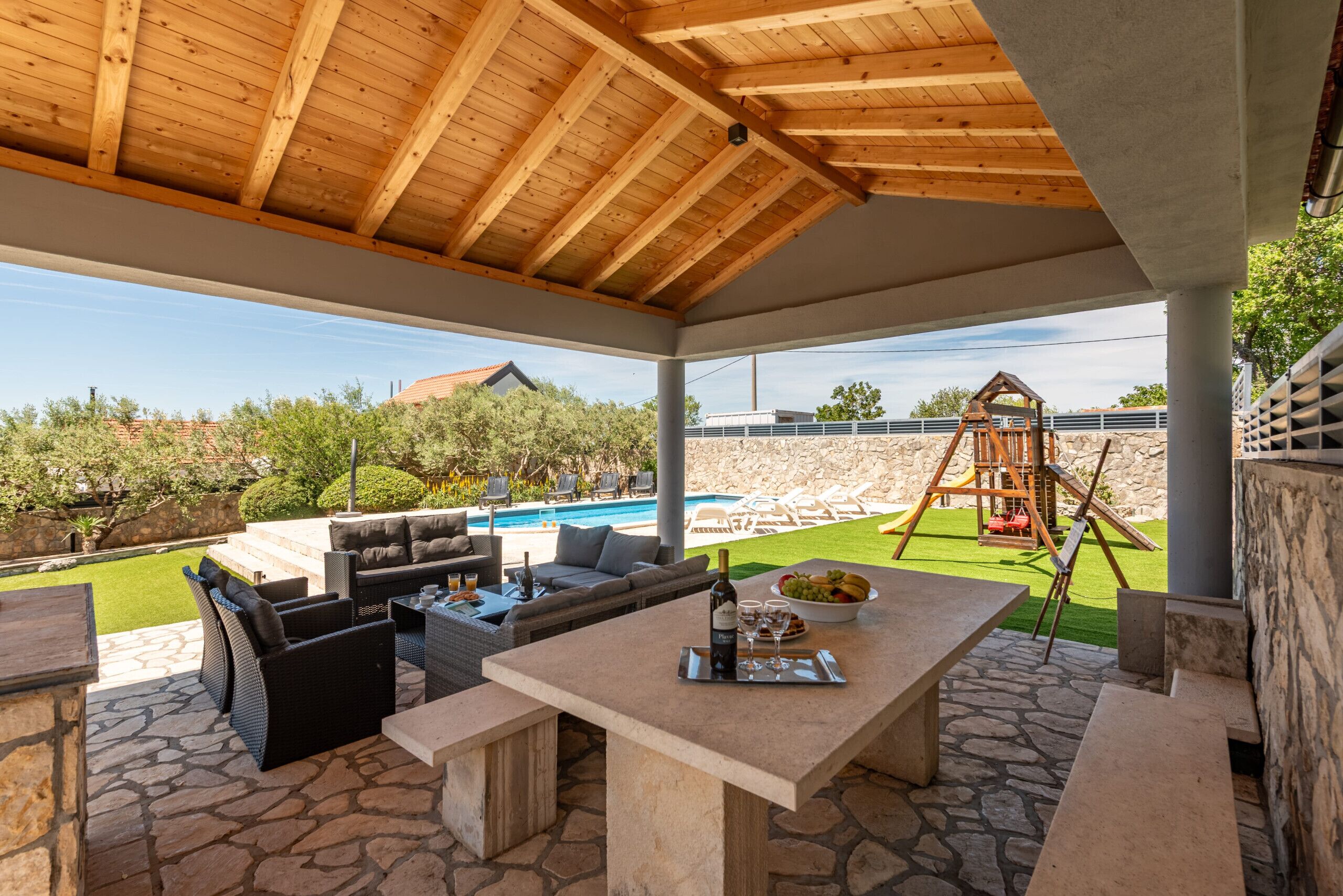 Eine Außenpatio mit Steinböden, Holzboden, einem Esstisch mit Flasche und Gläsern sowie Blick auf ein Schwimmbad und eine Spielanlage für Kinder.