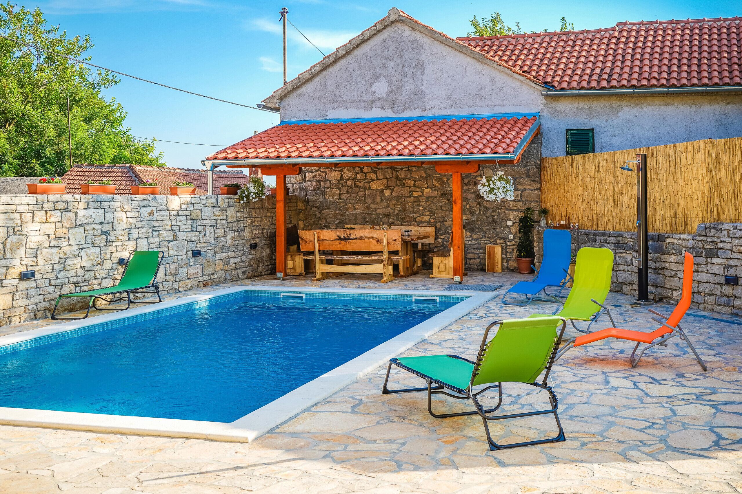 A swimming pool with lounge chairs, a stone wall, and a building with a tiled roof.