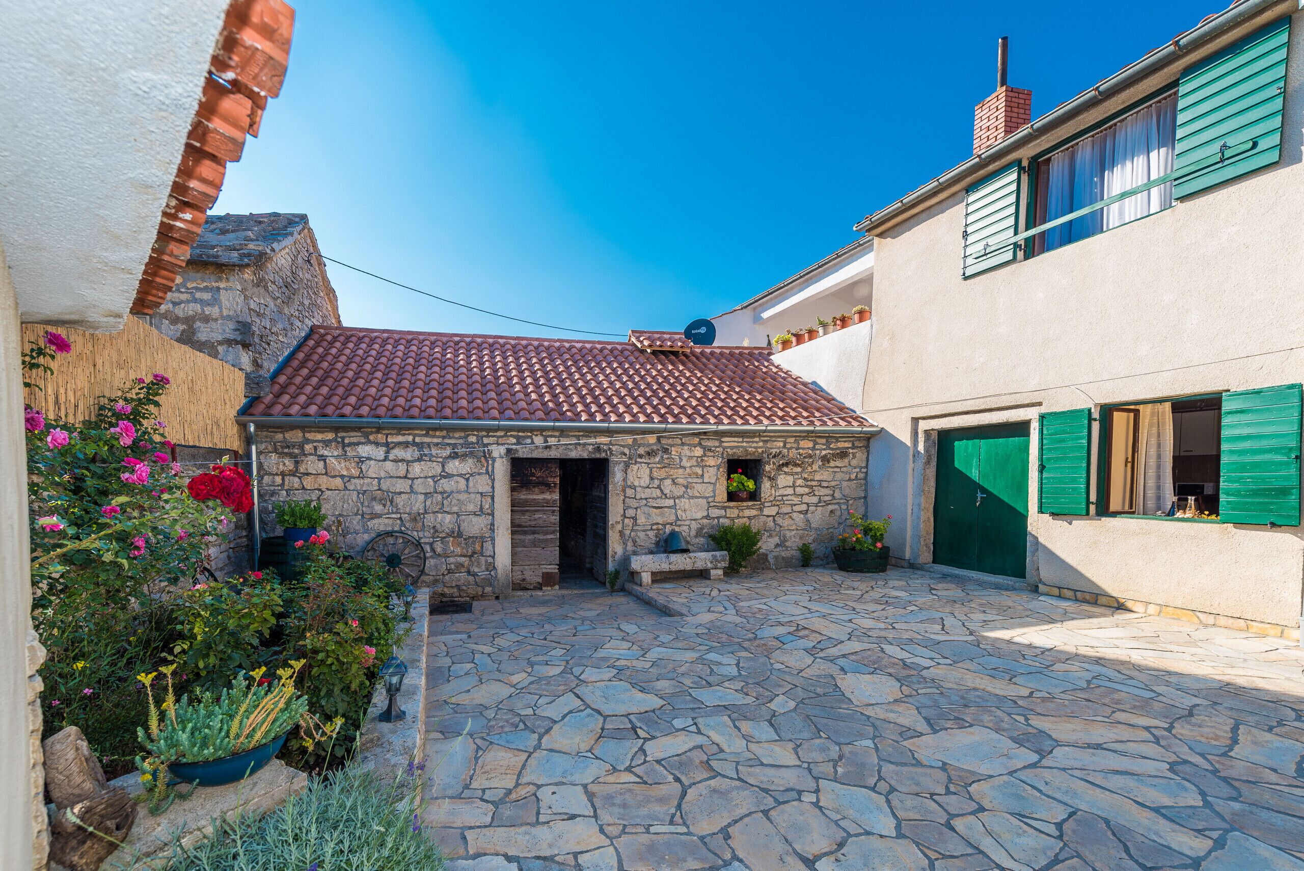 A stone building with a tiled roof, a stone courtyard, and a small garden with flowers.