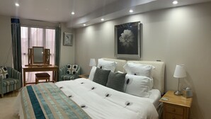 A hotel room with a large bed, bedside lamps, a desk with a mirror, and a framed picture on the wall. - King Canute Apartment - Crown Lettings (Chichester)