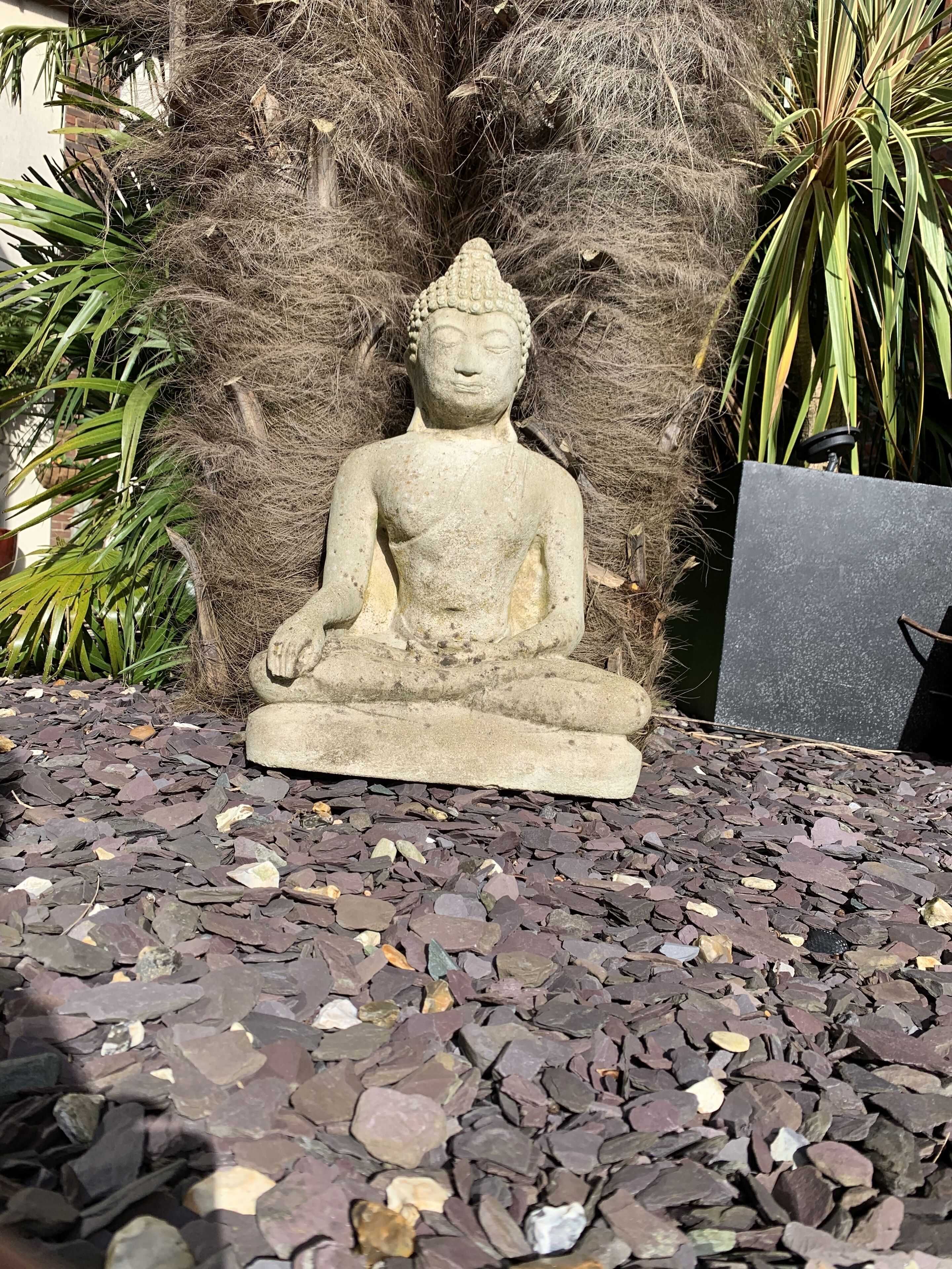 Una estatua de Buda de piedra sobre un lecho de guijarros multicolores, rodeada de hierba seca y plantas verdes.