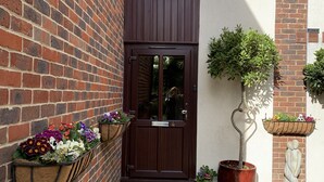 A brick building with a dark wooden door, flower boxes on the wall, and a potted tree. - King Canute Apartment - Crown Lettings (Chichester)