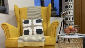 A mustard-colored armchair with a patterned throw blanket, a small round table with a metal wire base and a potted plant, and a window with a view of the outside. - King Canute Apartment - Crown Lettings (Chichester)