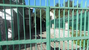 Een groen metalen hek met verticale spijlen, dat leidt naar een verhard gebied met een gebouw op de achtergrond.
