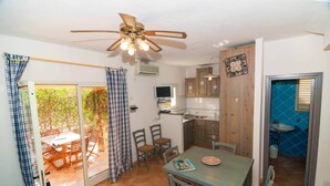 Una habitación con una mesa de comedor, sillas, ventilador de techo, zona de cocina con armarios, un lavamanos y una ventana con cortinas.